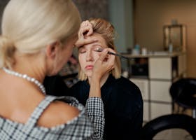 Crop anonymous beauty master doing makeup to female model with closed eyes using applicator in salon