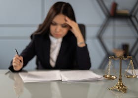 A fatigued female lawyer reviewing documents with a justice scale nearby, emphasizing stress in legal work.
